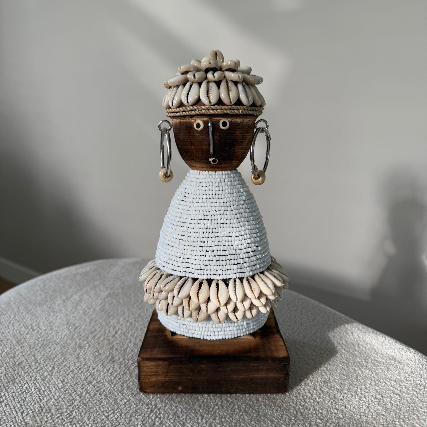Handgemaakte Namji Doll houten pop van circa 25 cm, versierd met witte glaskralen, kauri schelpen en oorbellen, uniek cultureel decoratiestuk
